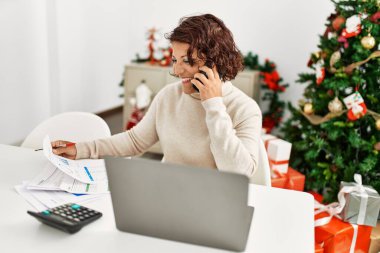 Orta yaşlı İspanyol kadın dizüstü bilgisayar kullanıyor ve akıllı telefonla konuşuyor. Yılbaşı ağacının yanında oturuyor..