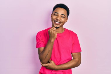 Casua tişörtü giyen genç Afro-Amerikan adam kameraya kendinden emin bakıyor kollarını kavuşturmuş ve çenesini kaldırmış gülümsüyor. Olumlu düşünmek. 