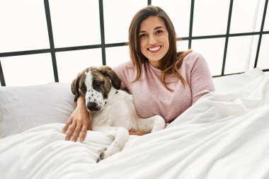 Genç kadın gülümsüyor. Kendine güvenen, sarılan köpek yatakta oturuyor.