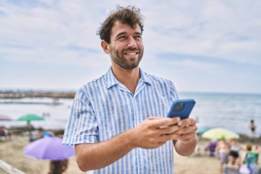 Genç İspanyol adam sahilde akıllı telefon kullanarak mutlu bir şekilde gülümsüyor.