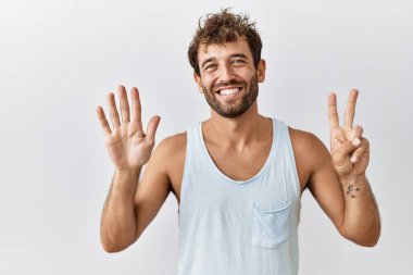 Genç yakışıklı adam izole edilmiş arka planda duruyor ve yedinci parmaklarını gösterip işaret ederken kendinden emin ve mutlu gülümsüyor.. 