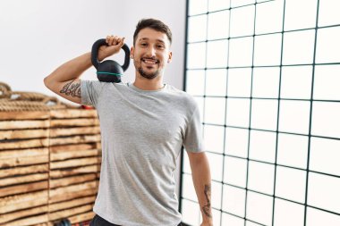 Spor kıyafetleri giyen genç yakışıklı adam halterler kullanıyor. Pozitif görünüyor ve mutlu görünüyor. Kendine güvenen bir gülümsemeyle dişlerini gösteriyor. 