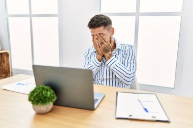 Young handsome man with beard working at the office using computer laptop with sad expression covering face with hands while crying. depression concept. 