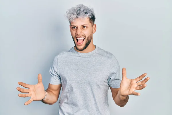 Young hispanic man with modern dyed hair wearing casual grey t shirt crazy and mad shouting and yelling with aggressive expression and arms raised. frustration concept.