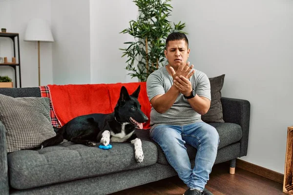 Young latin man and dog sitting on the sofa at home suffering pain on hands and fingers, arthritis inflammation