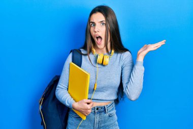 Genç esmer kız öğrenci çantası ve kulaklık takarak zaferi kutluyor mutlu bir gülümsemeyle ve ellerini kaldırarak kazanan ifadeyle. 