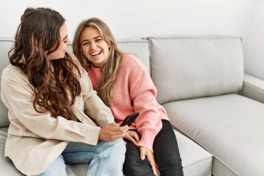 Genç çift evde koltukta oturmuş akıllı telefon kullanarak mutlu bir şekilde gülümsüyor..