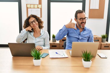 Orta yaşlı İspanyol kadın ve erkek ofiste dizüstü bilgisayarlarıyla oturup gülümsüyor telefonla el ve parmaklarıyla telefonla konuşuyor gibi davranıyorlar. iletişim kavramları. 