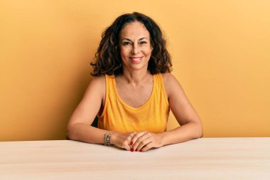 Masada mutlu ve havalı bir gülümsemeyle oturan, gündelik giysiler giyen orta yaşlı güzel bir kadın. şanslı kişi. 