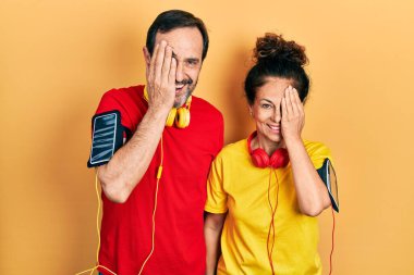 Orta yaşlı bir çift İspanyol kadın ve erkek spor giysisi ve kol bandı takıyor. Bir gözünü eliyle kapatıyor. Yüzünde kendine güvenen bir gülümseme ve sürpriz bir duygu var.. 