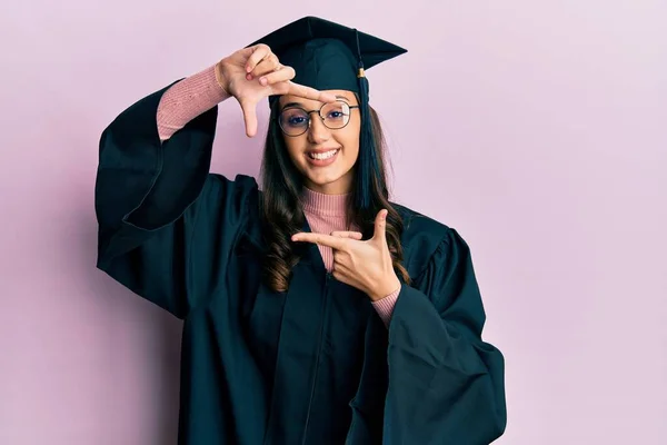 卒業キャップと式のローブを身に着けている若いヒスパニック系の女性は幸せな顔で手や指でフレームを作り笑顔 創造性と写真の概念 — ストック写真