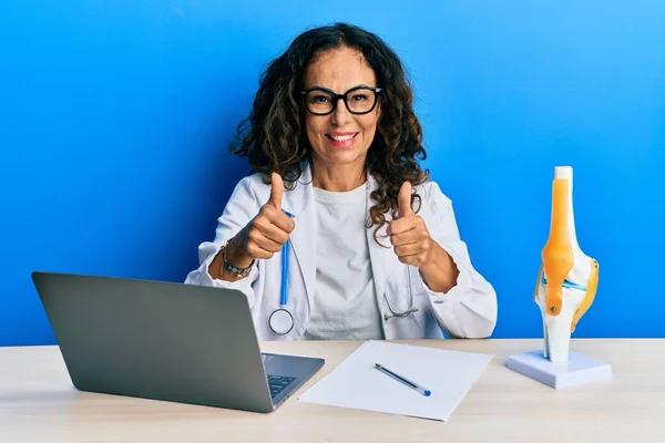 Beautiful middle age woman doctor at orthopedic clinic success sign doing positive gesture with hand, thumbs up smiling and happy. cheerful expression and winner gesture.