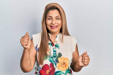 Orta yaşlı İspanyol kadın gündelik giysiler giyip çok mutlu ve heyecanlı bir şekilde kollarını kaldırıp, gülümseyip, başarı için haykırarak kazanan bir jest yapıyor. kutlama konsepti. 