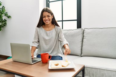 Genç Latin kadın kahvaltısını yapıyor. Evdeki kanepede oturuyor.