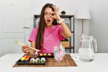 Genç esmer kadın yemek çubuklarıyla suşi yiyor. Şaşırmış yüz ve gözlerle parmaklarından bakıyor. İnançsız ifade. 