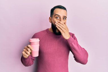 Sakallı genç adam bir fincan kahve içerek ağzını eliyle kapatıyor, şok geçiriyor ve hata yapmaktan korkuyor. şaşırmış ifade 