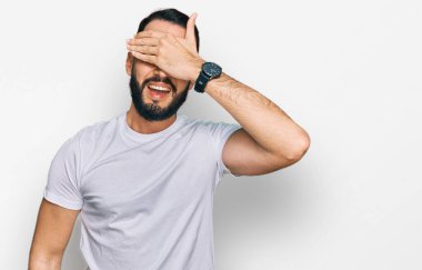 Sakallı, sıradan beyaz tişörtlü, gülümseyen ve elleri yüzünde, şaşırtmak için gözlerini kapatan genç bir adam. kör kavram. 