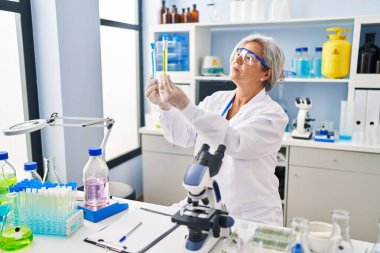 Laboratuvarda test tüpleri arayan orta yaşlı bir kadın bilim adamı üniforması giyiyor.