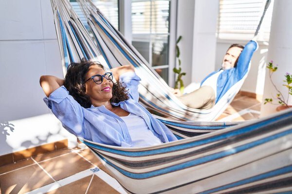 Middle age hispanic couple relaxed with hands on head lying on hammock at terrace
