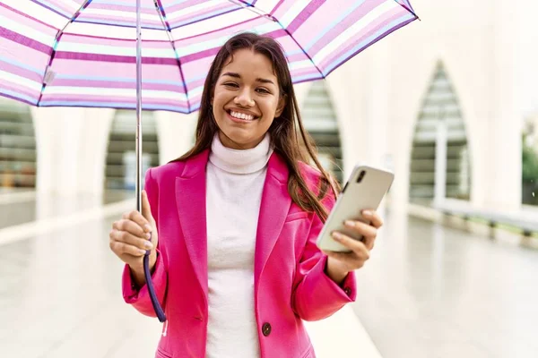Young latin woman using smartphone holding umbrella at street