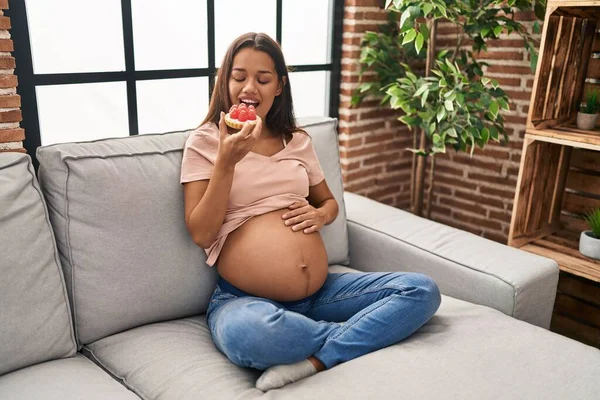 若いですラテン女性妊娠食べるケーキ座っています上のソファにホーム — ストック写真