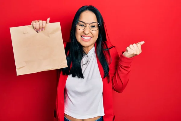 Beautiful hispanic woman with nose piercing holding take away paper bag screaming proud, celebrating victory and success very excited with raised arm