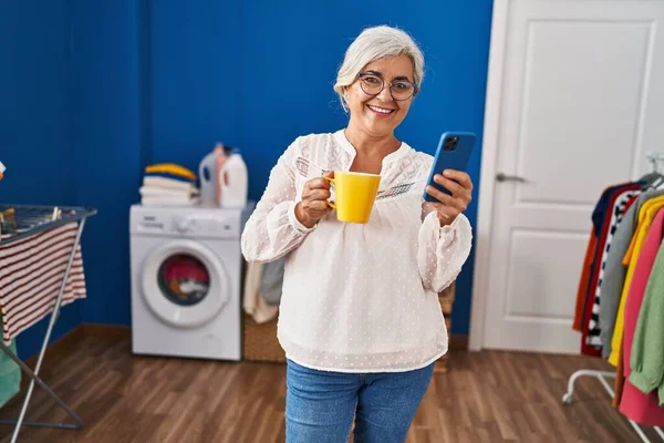 Medelålders Kvinna Med Smartphone Dricka Kaffe Väntar Tvättmaskin Tvättstugan — Stockfoto