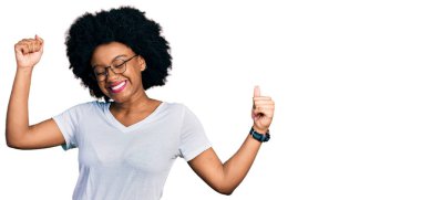 Genç Afrikalı Amerikalı kadın, günlük beyaz tişört giyip mutlu ve neşeli dans ediyor, gülümsüyor, rahat ve kendinden emin bir şekilde müzik dinliyor. 