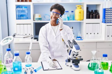 Bilim adamı üniforması giyen Afrikalı Amerikalı kadın. Akıllı telefondan konuşuyor. Laboratuvarda panoya yazı yazıyor.