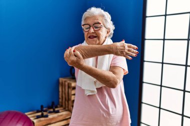 Spor kıyafetleri giyen yaşlı gri saçlı kadın spor merkezinde kendine gülerek duruyor.