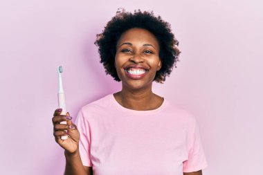 Elektrikli diş fırçası tutan Afro-Amerikalı genç bir kadın. Pozitif ve mutlu görünüyor. Kendine güvenen bir gülümsemeyle dişlerini gösteriyor. 