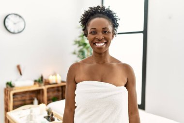 Young african american woman wearing towel standing at beauty center looking positive and happy standing and smiling with a confident smile showing teeth 