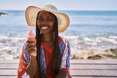 Young african american woman eating ice cream sitting on the bench at the beach.