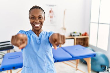 Saçları örgülü siyah bir kadın, ağrı kesici kliniğinde çalışıyor. Seni ve kamerayı işaret ediyor. 