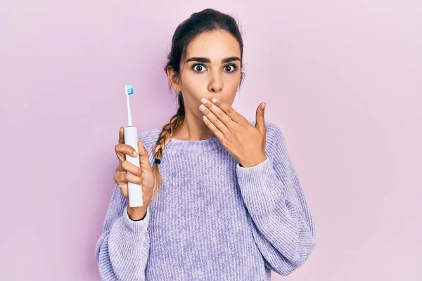 Young hispanic girl holding electric toothbrush covering mouth with hand, shocked and afraid for mistake. surprised expression