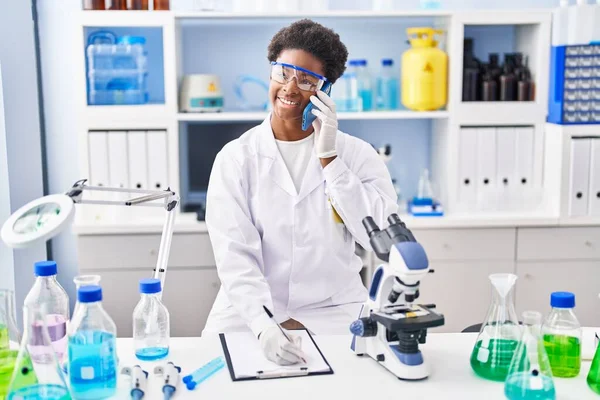 Bilim adamı üniforması giyen Afrikalı Amerikalı kadın. Akıllı telefondan konuşuyor. Laboratuvarda panoya yazı yazıyor.