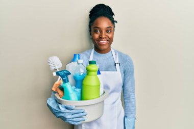 Saçları örgülü, önlük takan, temizlik ürünleri tutan, pozitif görünen, gülümseyen, kendine güvenen, dişlerini gösteren Afro-Amerikan bir kadın. 