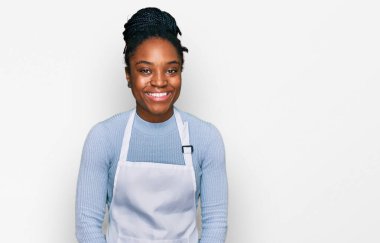 Önlük takan Afro-Amerikalı genç bir kadın pozitif ve mutlu görünüyor ve kendine güvenen bir gülümsemeyle dişlerini gösteriyor. 