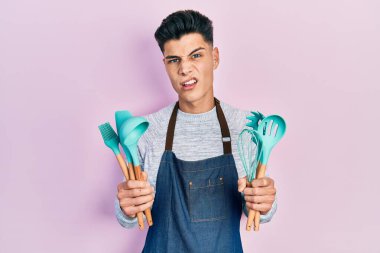 Profesyonel fırıncı önlüğü giyen genç İspanyol adam yemek pişirme aletlerini elinde tutuyor. Hiçbir şeyden habersiz ve kafası karışık. şüphe kavramı. 