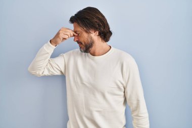 Mavi arka plan üzerine sıradan süveter giyen yakışıklı orta yaşlı adam yorgun burnu ve gözleri ovuşturup yorgun ve baş ağrısı çekiyor. Stres ve hüsran kavramı. 