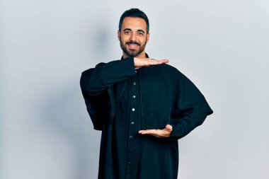 Sakallı, katolik rahip cüppesi giyen, elleri büyük ve büyük bir işaret gösteren yakışıklı bir İspanyol. Ölçü sembolü. Kameraya bakıp gülümsüyor. ölçüm kavramı. 