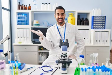 Laboratuvarda çalışan, sakallı, neşeli gülümseyen, elinde avucuyla kameraya bakan genç İspanyol bir adam.. 