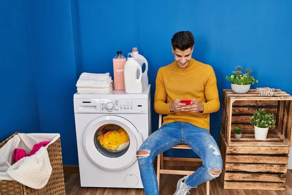 Young Hispanic Man Using Smartphone Waiting Washing Machine Laundry —  Fotos de Stock
