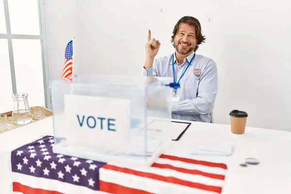 一个英俊的中年男人坐在投票站里 脸上挂着大大的笑容 手指手画脚地望着摄像机的侧面 — 图库照片