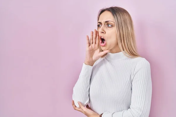 ピンクの背景に白いセーターを着た若いブロンドの女性が叫び 大声で口の中で手で横に叫んでいます コミュニケーションの概念 — ストック写真