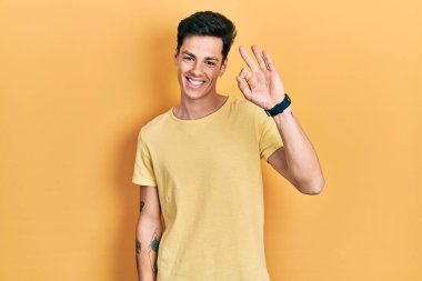 Günlük sarı tişört giyen genç İspanyol adam gülümsüyor ve el ve parmaklarıyla 