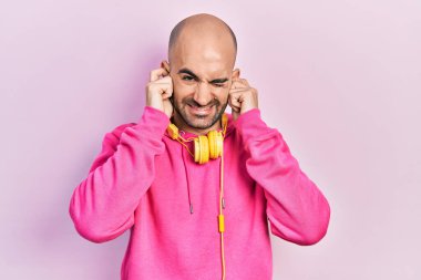 Spor kıyafetleri giyen ve kulaklık takan genç kel bir adam yüksek sesli müzik sesi için parmaklarıyla kulaklarını kapatıyor. sağır kavramı. 