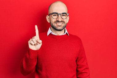 Genç kel bir adam günlük kıyafetler ve gözlük takıyor. Bir numaralı parmağını gösteriyor ve işaret ediyor. Kendinden emin ve mutlu bir şekilde gülümsüyor.. 