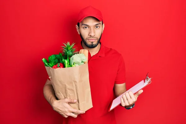 Hispanic man with beard wearing courier uniform with groceries from supermarket and clipboard relaxed with serious expression on face. simple and natural looking at the camera.