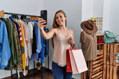 Genç beyaz kadın müşteri elbise mağazasında akıllı telefon alışverişinde selfie çekiyor.